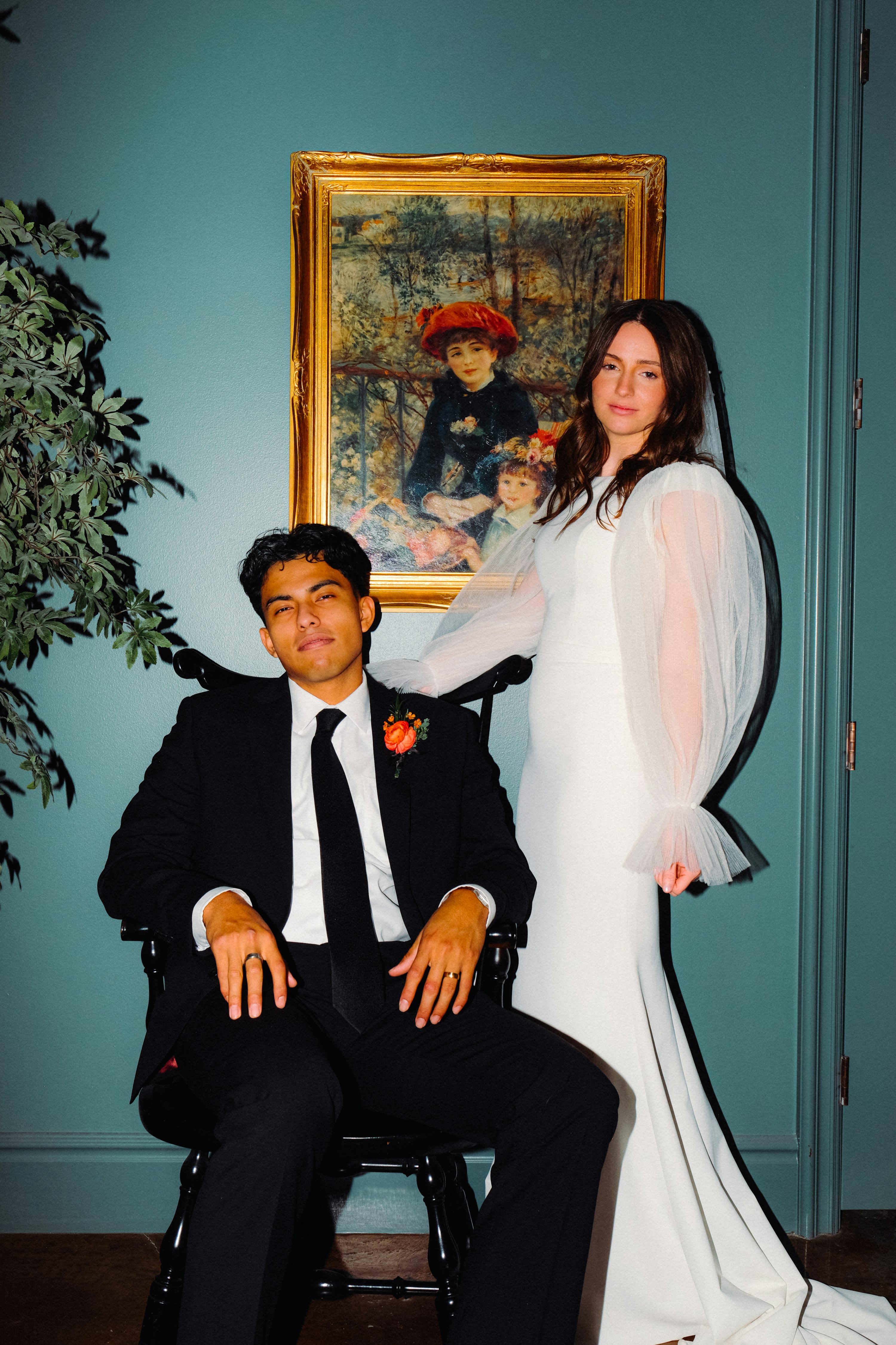 full shot of a bride and groom posing for a wedding portrait. the bride is wearing a long, white, formfitting gown with sheer, puffed sleeves. the groom is in a black suit and tie. they are seated in front of a large painting in a gold frame. the background is a tealcolored wall.