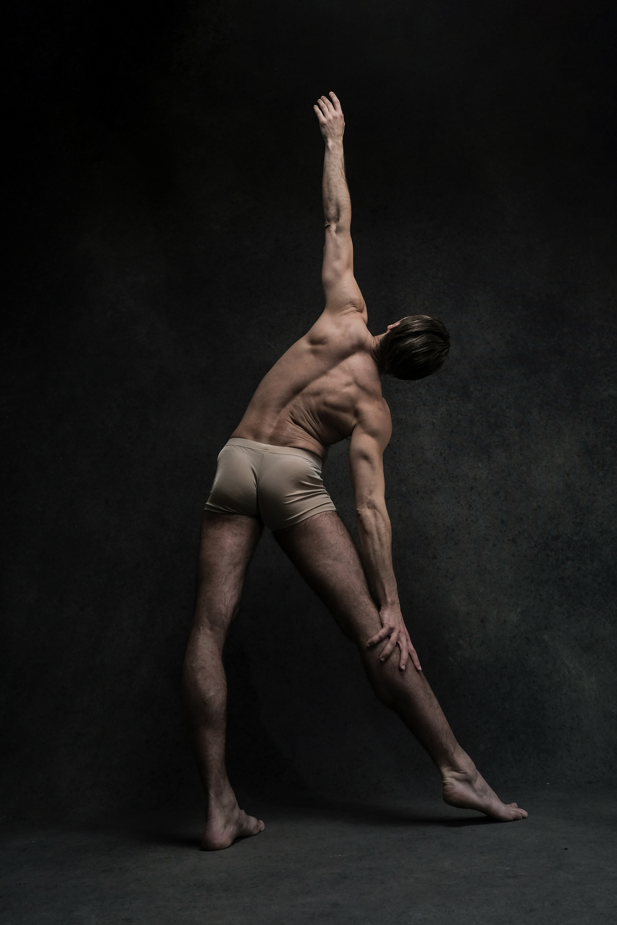 a male ballet dancer is captured midpose against a dark, textured background. the spotlight illuminates his muscular back and the elegant lines of his body as he stretches with one arm raised and the other reaching towards his ankle.  his posture conveys both strength and flexibility, highlighting the athleticism and artistry of ballet.
