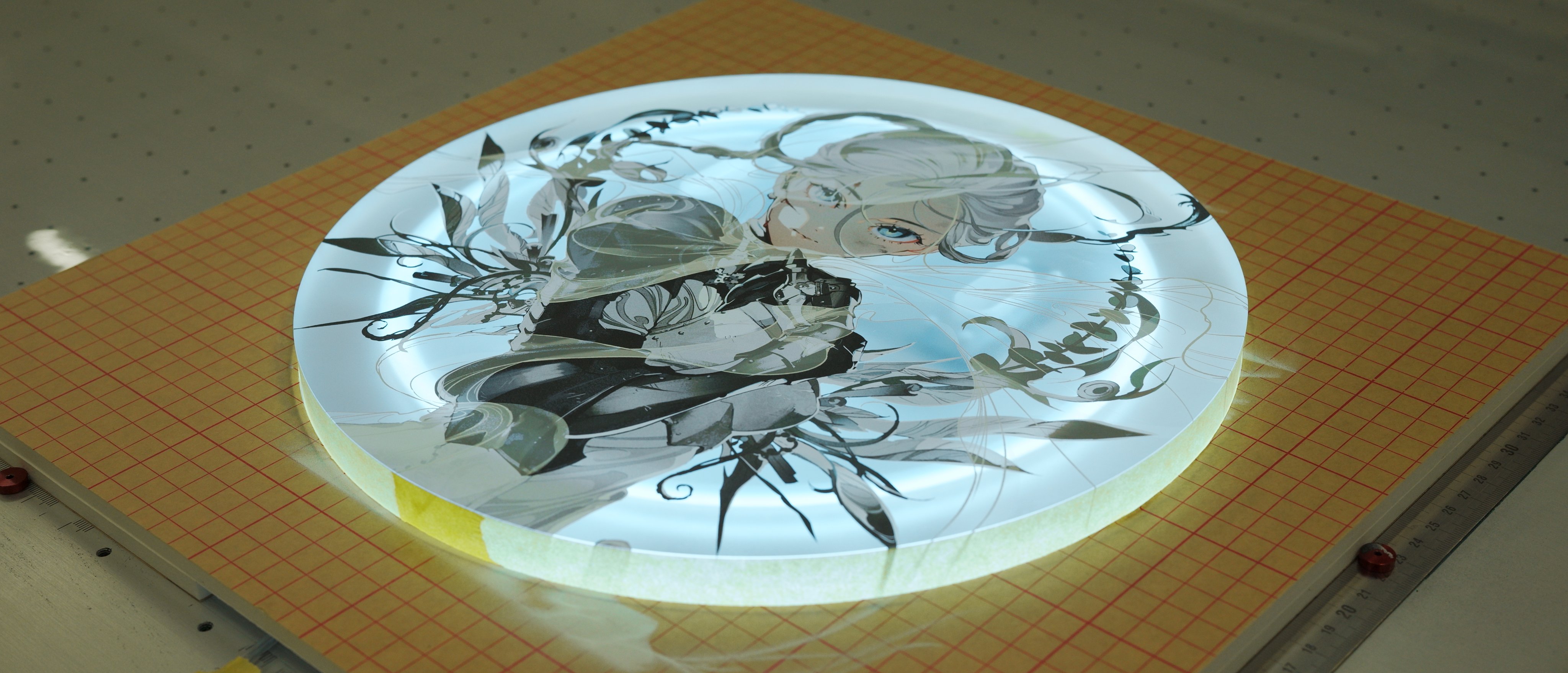 a round, backlit print of an animestyle illustration is displayed on a table. the illustration features a woman with white hair and a black and white outfit. the print is illuminated from behind, creating a soft glow around the image. the table is covered with a gridded cutting mat, suggesting a studio or workshop setting.