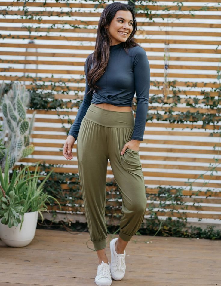 a woman is standing on a patio, posing for the camera. she is wearing a casual outfit consisting of a longsleeved, dark gray crop top, olive green harem pants, and white sneakers. the background features a wooden fence partially covered with greenery and a potted plant. the lighting is natural and the perspective is at eye level.