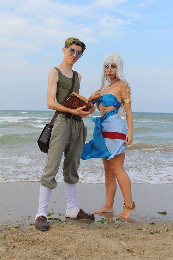 two cosplayers dressed as milo thatch and princess kida from the movie atlantis: the lost empire are standing on a sandy beach in front of the ocean. milo is holding a book and wearing glasses, a tank top, baggy pants, socks, and loafers. princess kida is wearing a bikini top, sarong, bare feet, and jewelry.