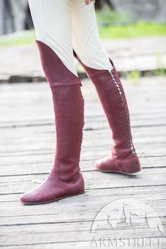the image shows a pair of women's kneehigh boots crafted from burgundy leather. the boots feature a distinctive side lace detail that extends from the ankle to the top of the boot. they are styled with a pair of creamcolored pants, offering a glimpse of the wearer's leg through a slit at the back.  the setting appears to be an outdoor wooden deck, bathed in natural daylight.