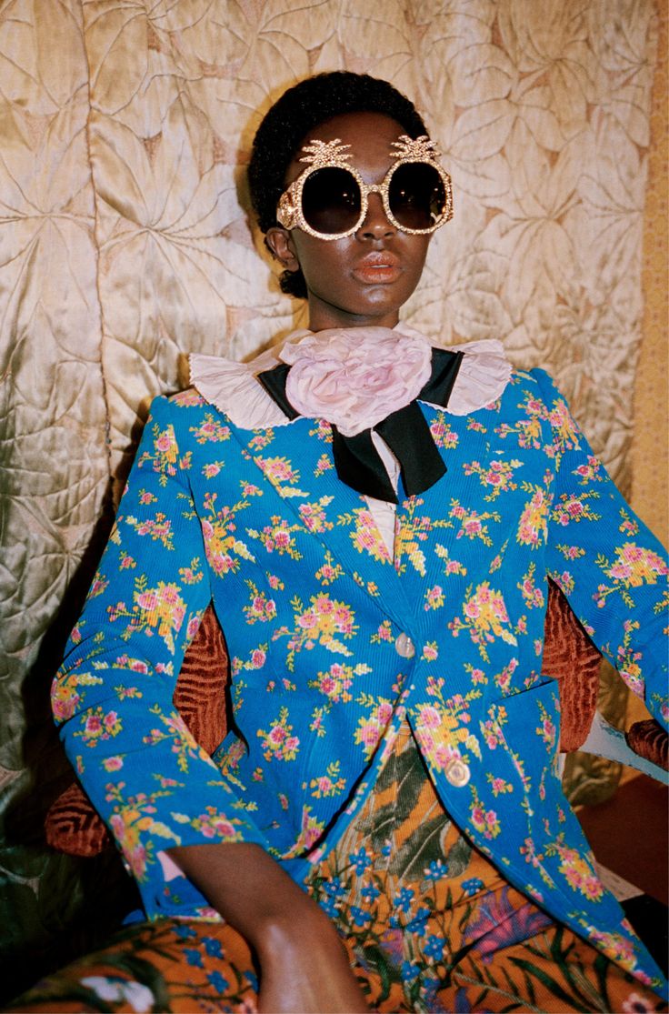 a closeup shot of a darkskinned model wearing a vibrant blue floral blazer and pants. the model is also adorned with large, round, gold sunglasses embellished with pineapples.  a light pink rose is fastened at her collar. the background features an ornate, quilted, gold wallpaper. the overall style is reminiscent of retro or vintage fashion. the image is brightly lit and the colors are bold and saturated.
