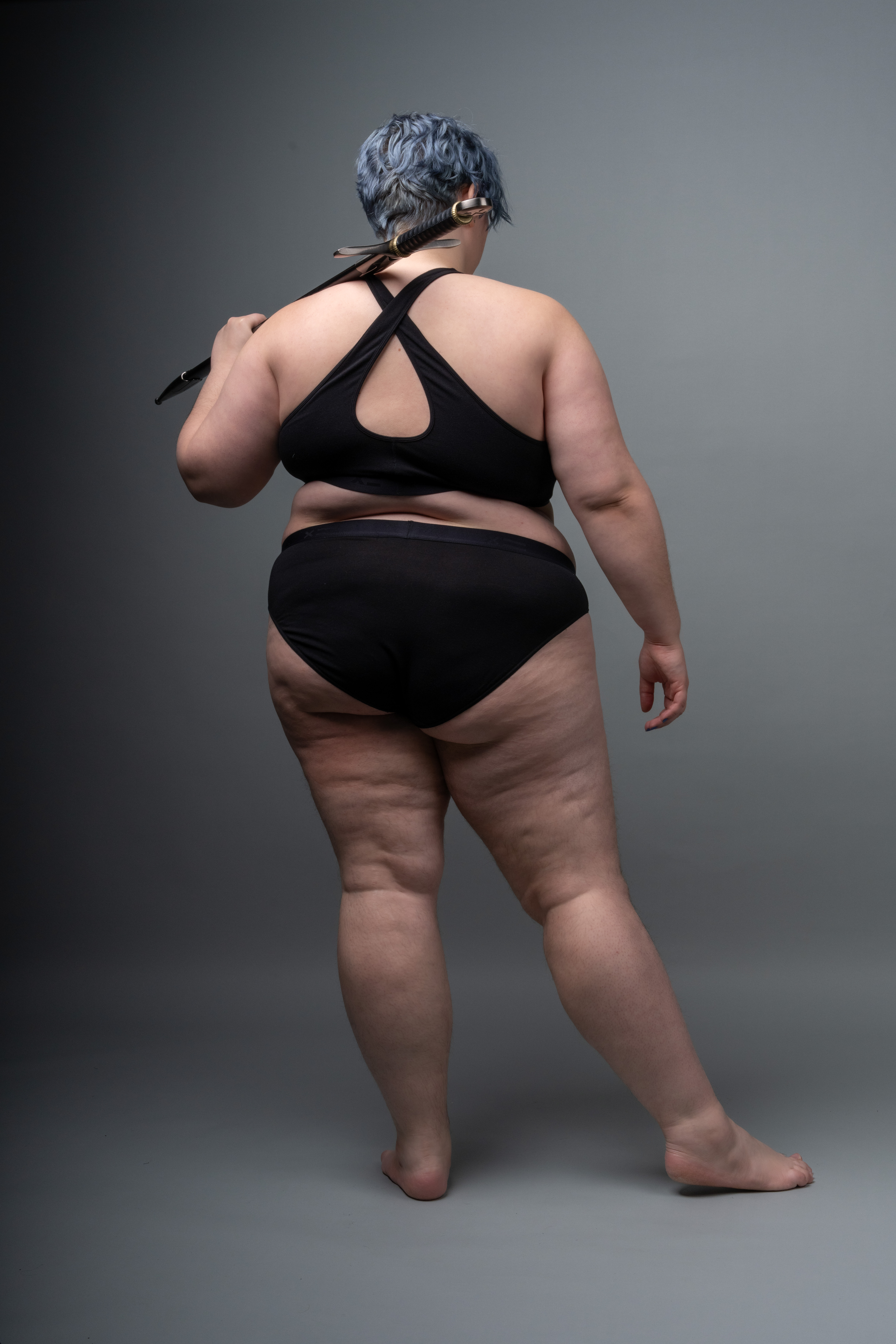 a plussize woman with blue hair stands with her back to the viewer, wearing a black bra and underwear. she holds a sword across her back. the background is a plain grey.