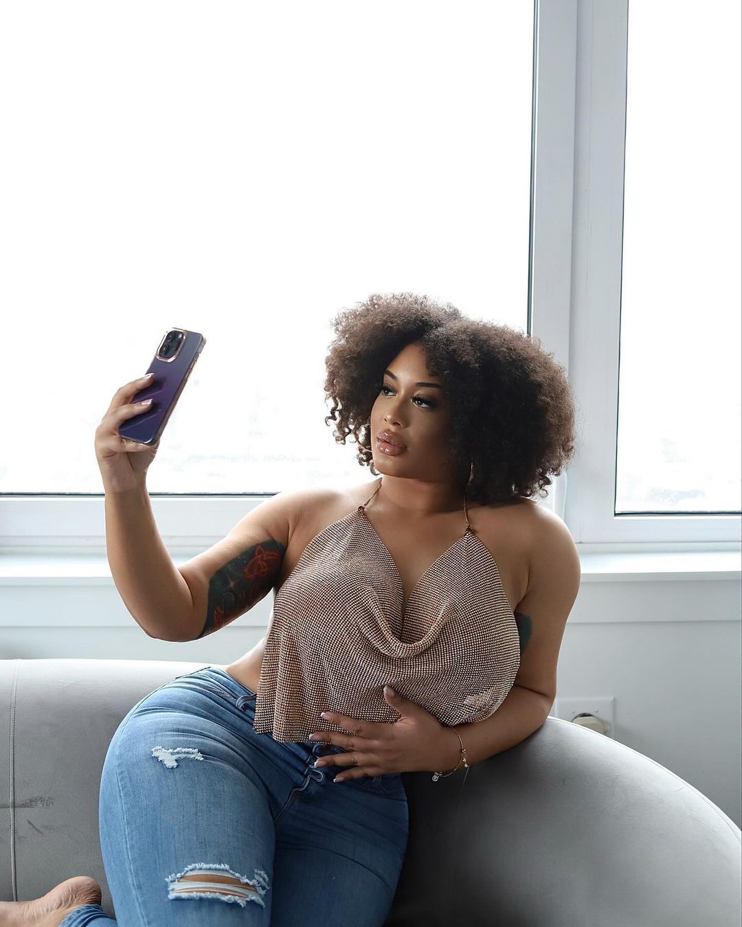 a young woman in a  jeans and a stylish top sits on a couch, looking at her phone. she has her hair down in  loose curls, her smile is full of happiness, and she is looking at the camera with a bright and joyful expression. she is casual but stylish and chic.