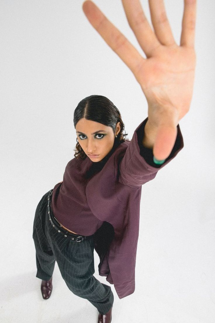a highangle, fullbody shot of a young woman wearing a maroon coat and striped trousers.  she is posing with one hand extended towards the camera, partially obscuring her face.  the background is plain white, and she is wearing dark shoes. her expression is serious. the image is styled and fashionoriented.