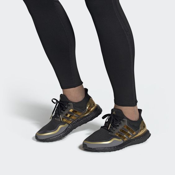 a person is wearing a pair of black and gold adidas ultraboost running shoes. the shoes have a black primeknit upper, a black boost midsole, and a black rubber outsole. the adidas logo on the side of the shoes is gold. the person is wearing black pants. the background is white.