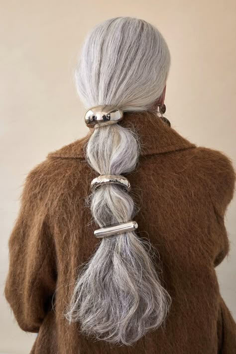 a woman with long, silver gray hair is wearing a brown fuzzy jacket. her hair is pulled back in a ponytail and adorned with three silver hair clips, each with a different shape. she looks classy and elegant.