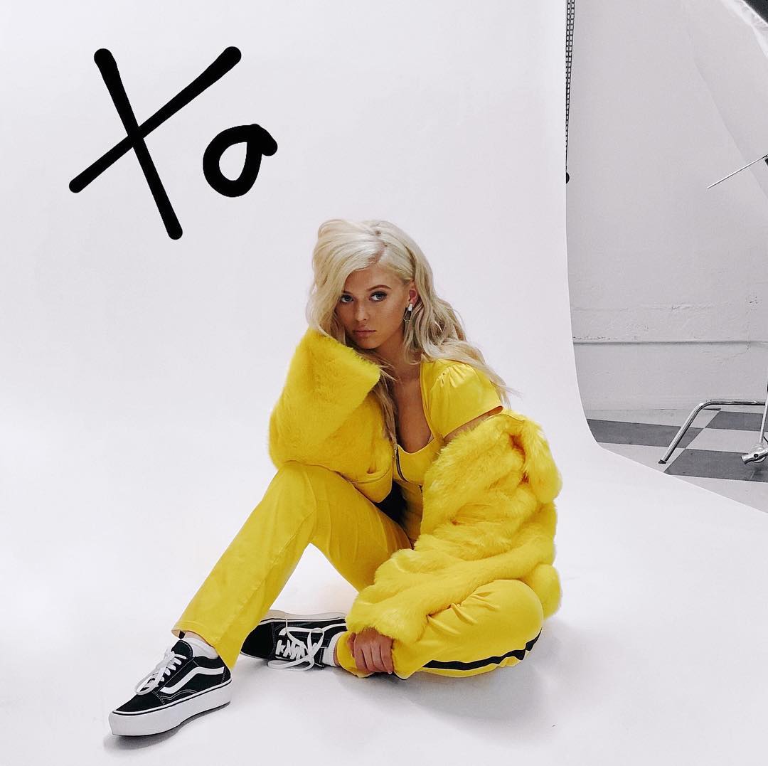 a woman with blonde hair is sitting on a white studio floor wearing a yellow outfit consisting of a top, trousers, and a fur coat. she is also wearing black and white vans platform sneakers and jewelry. a large black "xo" is drawn on the white background behind her. a photography light stand is visible in the background on the right.