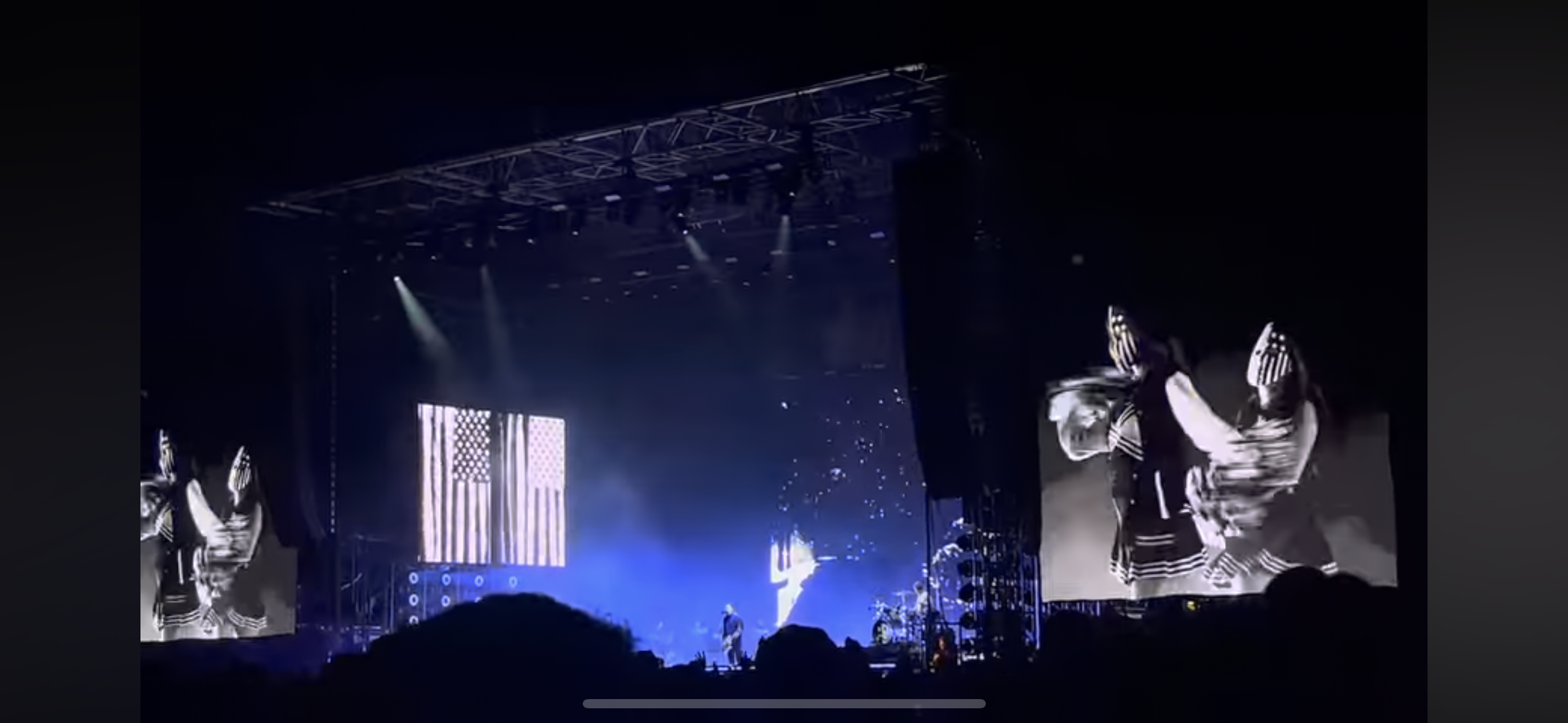 this is a photo taken at a concert. the stage is illuminated with bright stage lights and features a giant screen displaying an image of the american flag. in the background, the band is performing on stage, with a few people in the crowd and an empty space in front of the stage.