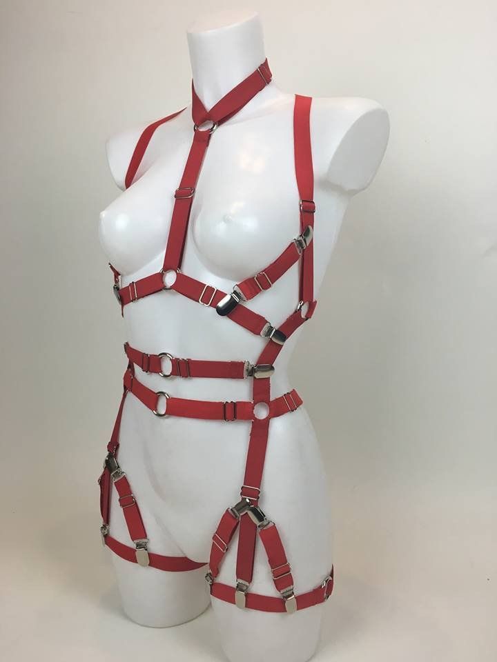 a red body harness with adjustable straps and metal hardware is displayed on a white mannequin against a white background. the harness includes a choker and various straps that wrap around the chest, waist, and thighs.