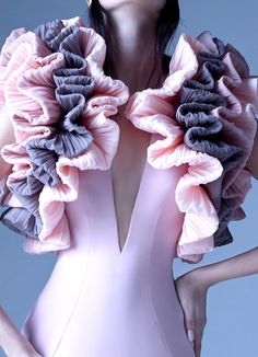 a woman is standing in a studio wearing a pink dress with gray ruffles on the shoulders. the dress has a deep vneckline. she is looking directly at the camera with a seductive look.  the image is taken close up with a shallow depth of field, which makes her appear more seductive. the lighting is soft and warm, which gives the image a romantic feel.  the ruffles on the shoulders are very elegant and add a touch of drama to the outfit.