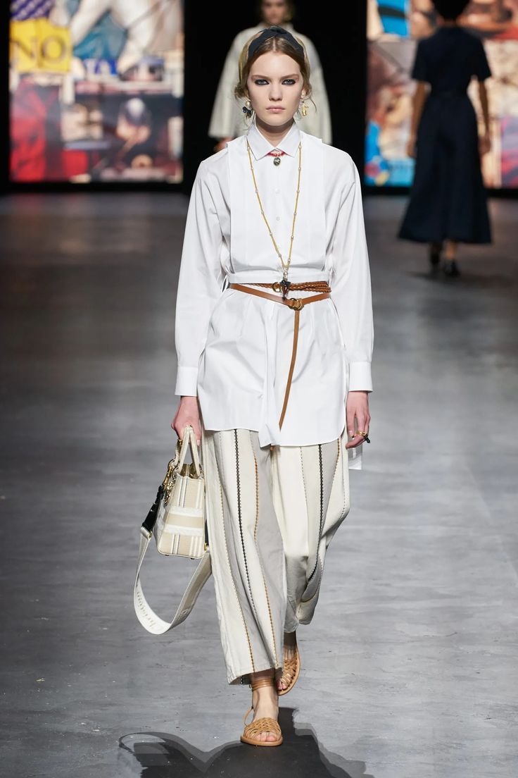 a female model walks down a runway wearing a white longsleeved shirt with a belt, white striped pants, sandals, and a bag. she has a headband on her head and jewelry around her neck and ears.  she looks straight at the camera as she poses on the runway.