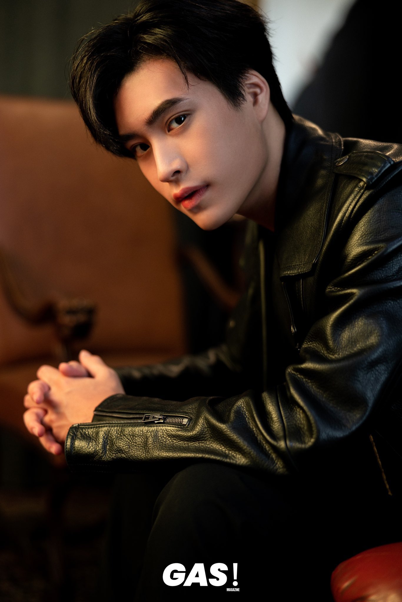 a young asian man with styled black hair poses for a fashion portrait. he wears a black leather jacket and sits with his hands clasped, looking directly at the camera with a neutral expression. the background is softly lit and out of focus, with warm brown and orange tones suggesting a comfortable indoor setting. the overall mood is stylish and approachable.
