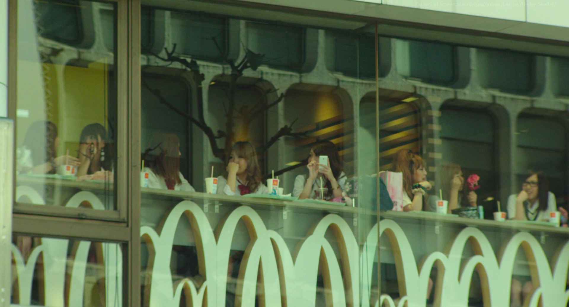 the image shows a group of young women eating and drinking at a mcdonald's restaurant. the photo is taken from outside the restaurant, looking in through a large window. the iconic golden arches of the mcdonald's logo are visible in the foreground, reflected in the window. the women are seated at a table and appear to be enjoying their meal. the restaurant is bright and airy, with natural light streaming in through the windows. the overall atmosphere is one of casual dining and socializing.