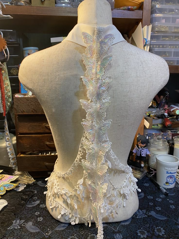 a mannequin is adorned with a piece of iridescent butterfly lace. the lace features a series of delicate butterflies arranged vertically, cascading down the back of the mannequin. it has a white collar and trim at the bottom, with decorative elements. the surrounding area is filled with craft supplies, suggesting a sewing or crafting environment.