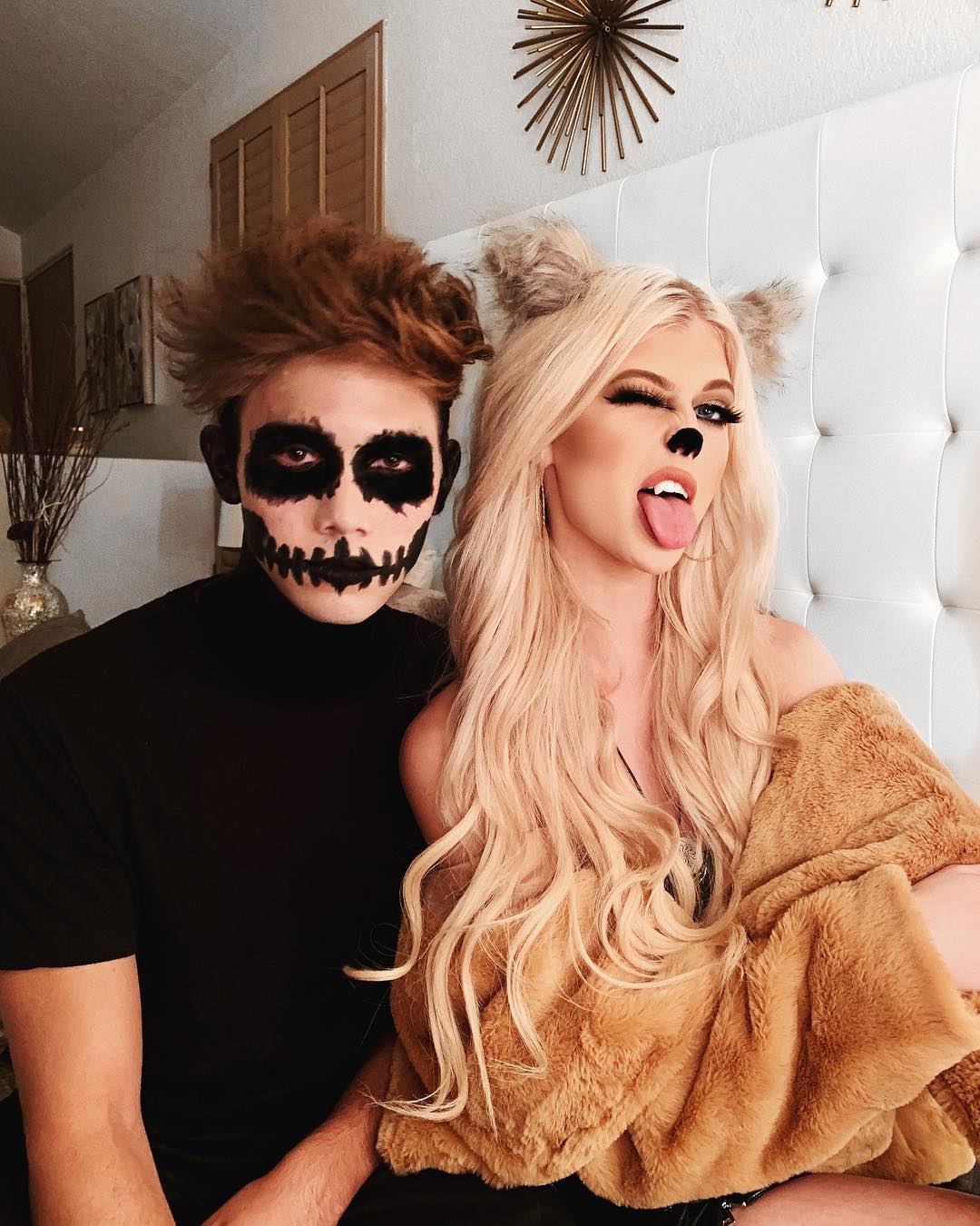 a couple in halloween costumes poses for a portrait in an interior setting. the man has skeleton makeup on his face and is wearing a black turtleneck. the woman has blonde hair, ears, a black nose, and is winking with her tongue out. she is wearing a brown fur coat.