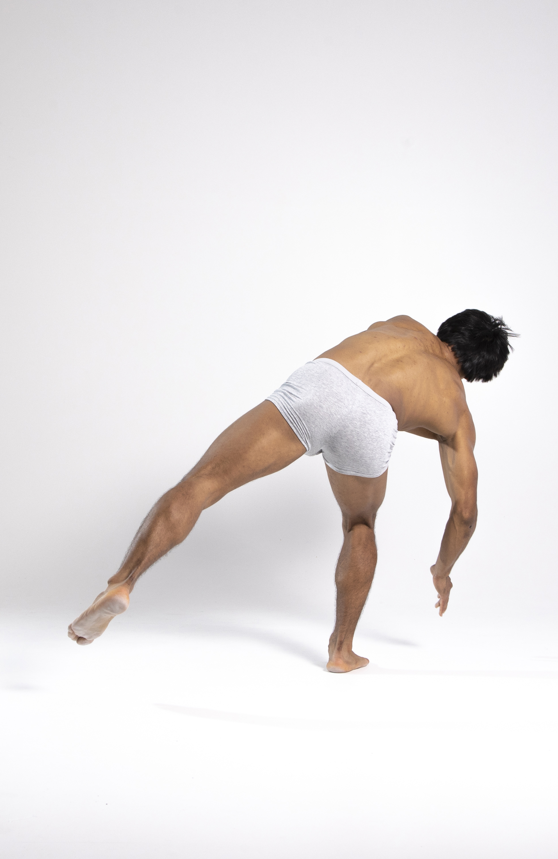 a man wearing gray underwear is captured in a dance pose against a plain white background. he is barefoot, with one leg extended behind him and the other supporting his weight. his torso is angled forward, and one arm is extended towards the floor. the image highlights his muscular physique and the flexibility required for dance.