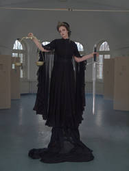 a full shot of a woman in a long, black, gothicstyle dress. she is holding a scale of justice in one hand and a sword in the other. the woman is standing in a large, empty room. the overall mood of the image is dark and mysterious.