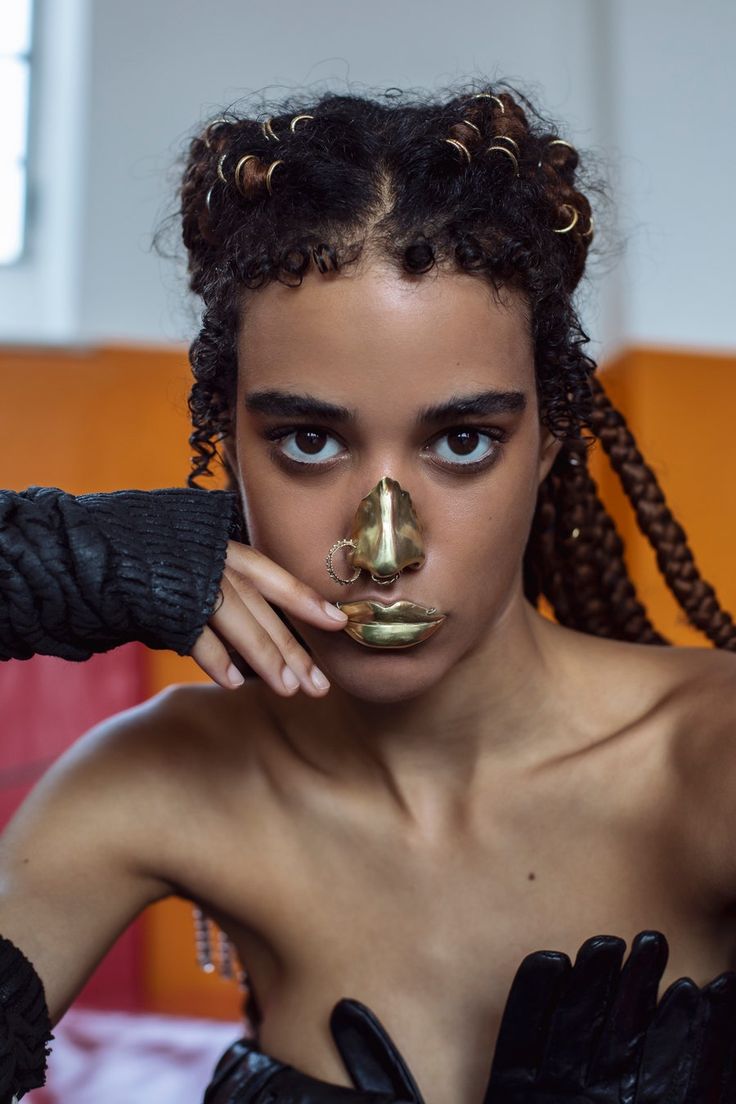 a close up portrait of a woman with black braided hair looking at the camera. she is wearing black gloves and has a gold nose ring, and lip ring.