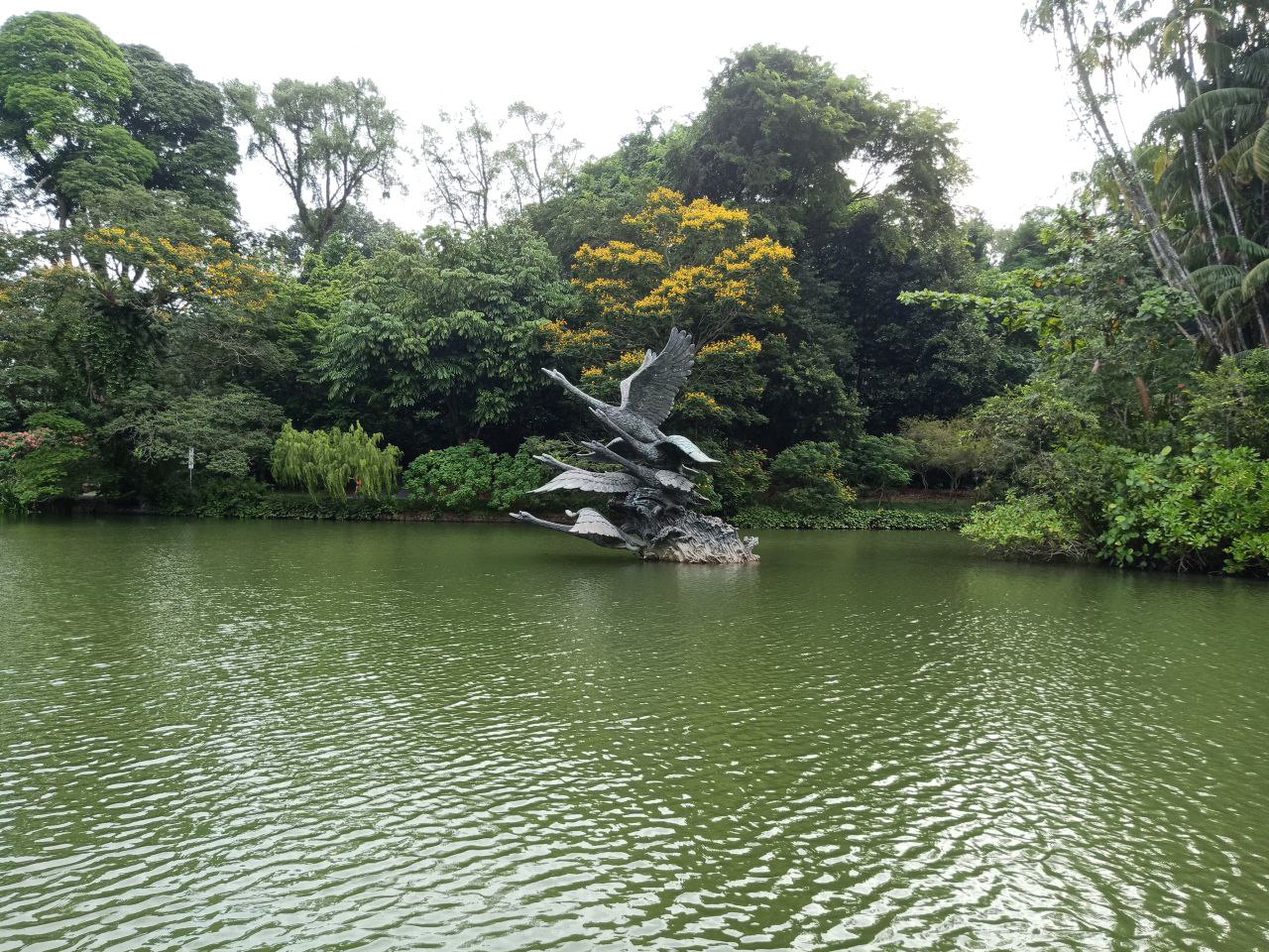 a beautiful view of a lake in a park, with lush green trees surrounding it. a swan sculpture stands in the middle of the lake, with its wings spread out, creating a serene and peaceful atmosphere. the water is calm and still, reflecting the surrounding trees and the clear blue sky. this is a beautiful and peaceful scene that evokes feelings of relaxation and tranquility.