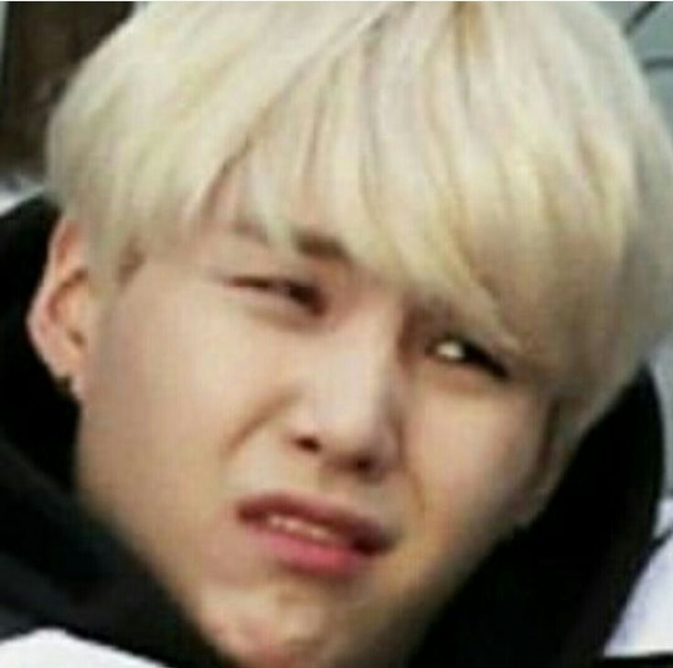 the image shows a closeup of an asian male with light blonde hair, squinting with a displeased facial expression. he appears to be wearing a hooded sweatshirt. the person is identified as suga (min yoongi) from the kpop group bts.