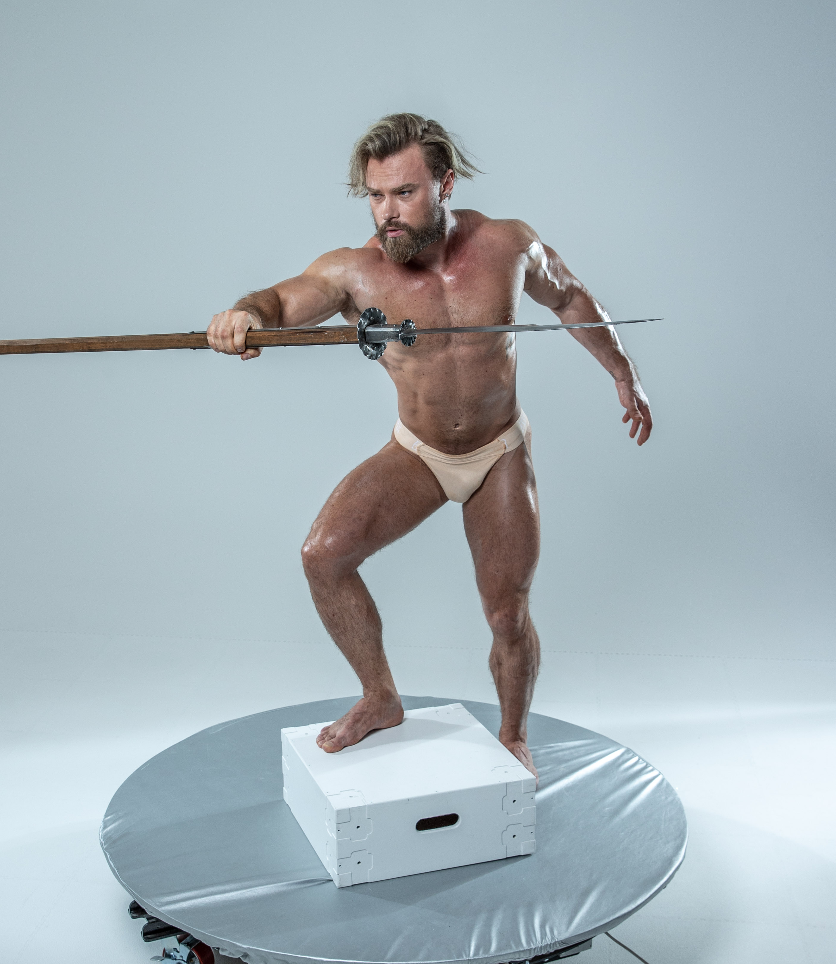 a muscular man with a beard and long blond hair pulled back from his face stands on a white platform in a photography studio, holding a spear level in front of himself. he is wearing only fleshtoned underwear. the background is light gray.