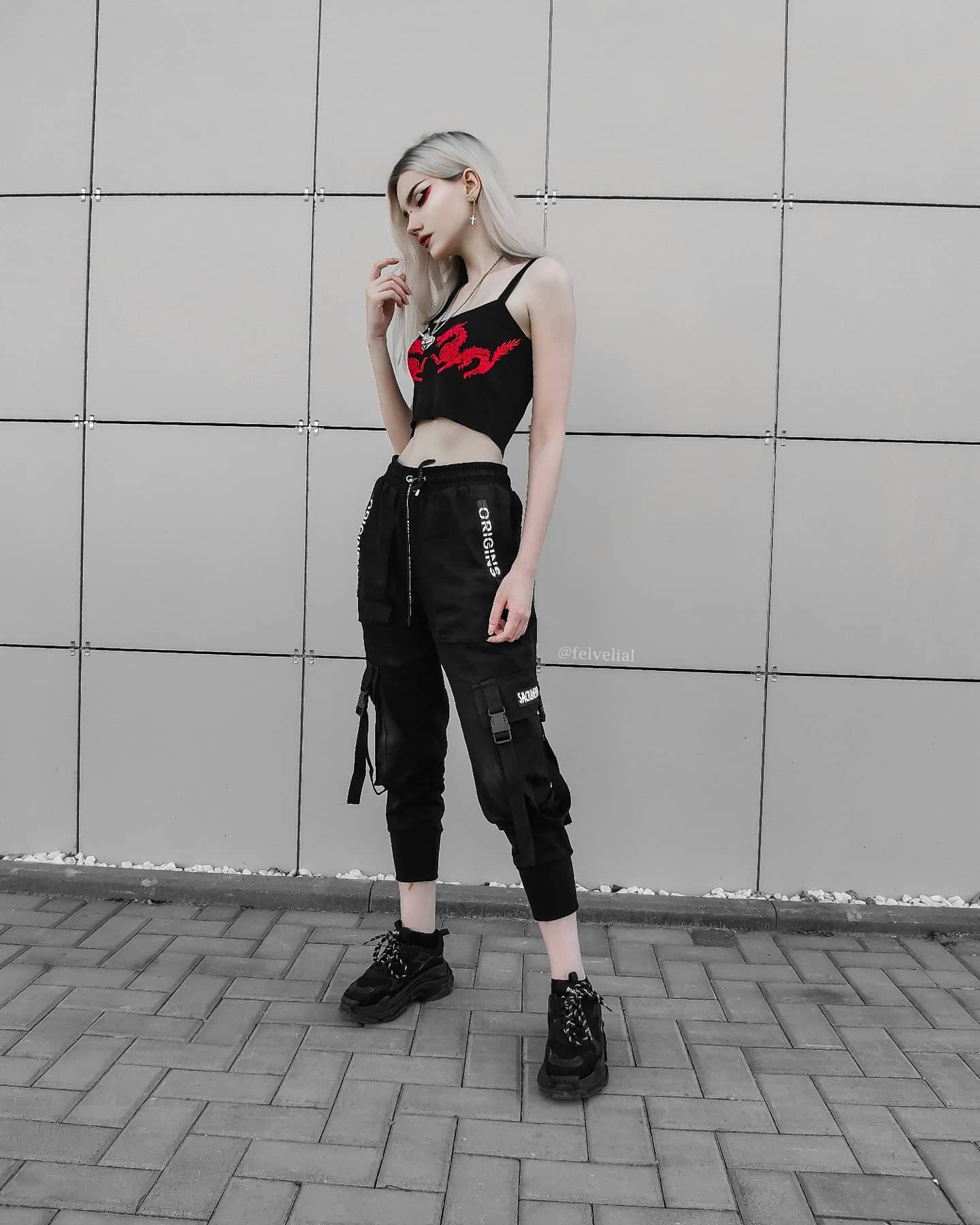 a stylish young woman with blonde hair is standing against a grey wall. she's wearing a black cropped top with a red dragon design, black cargo pants, and black chunky sneakers. her look is edgy and alternative, showcasing a blend of streetwear and goth aesthetics. the overall tone of the image is dark and moody.
