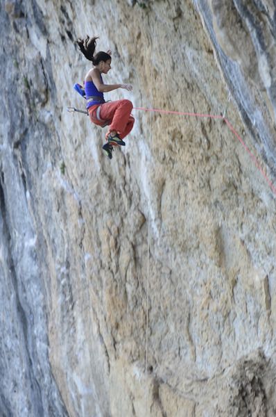 a female rock climber is captured midfall, suspended by her rope against a sheer rock face. her body is parallel to the ground, limbs outstretched, suggesting a controlled descent after losing her grip. the climber is the focal point, set against the textured, light gray and beige tones of the natural rock wall. natural daylight illuminates the scene, highlighting the climber's colorful attire and the raw beauty of the environment.