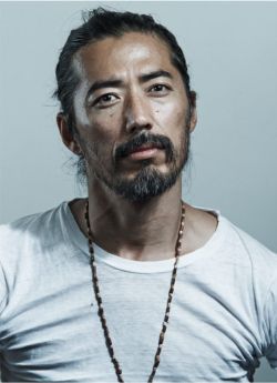 a portrait of an asian man with a beard and mustache wearing a white tshirt and a beaded necklace. his hair is tied back.