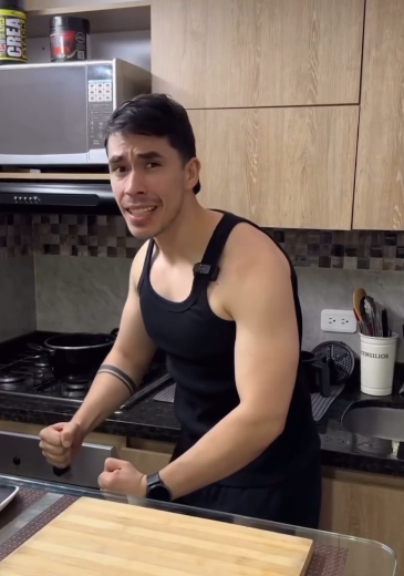 a man wearing a black tank top is standing in his kitchen, flexing his muscles. he has a microphone attached to his tank top. he has dark hair and a tattoo on his left arm. behind him are kitchen counters, a microwave, a container of protein powder, and some cooking supplies.