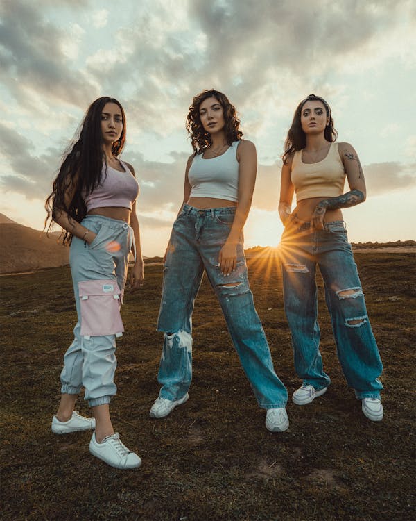 the image shows three young women standing in a grassy field at sunset.  two of the women wear baggy, ripped lightwash jeans and white sneakers; one wears light gray cargo pants with a pink pocket detail and white sneakers.  all three women wear crop tops – one pale pink, one white, one yellow. they are positioned in a slightly staggered line, facing the camera with confident expressions. the sunset creates a warm glow behind them, and mountains are visible in the distance. the overall aesthetic is casual and trendy, showcasing a youthful and stylish look.