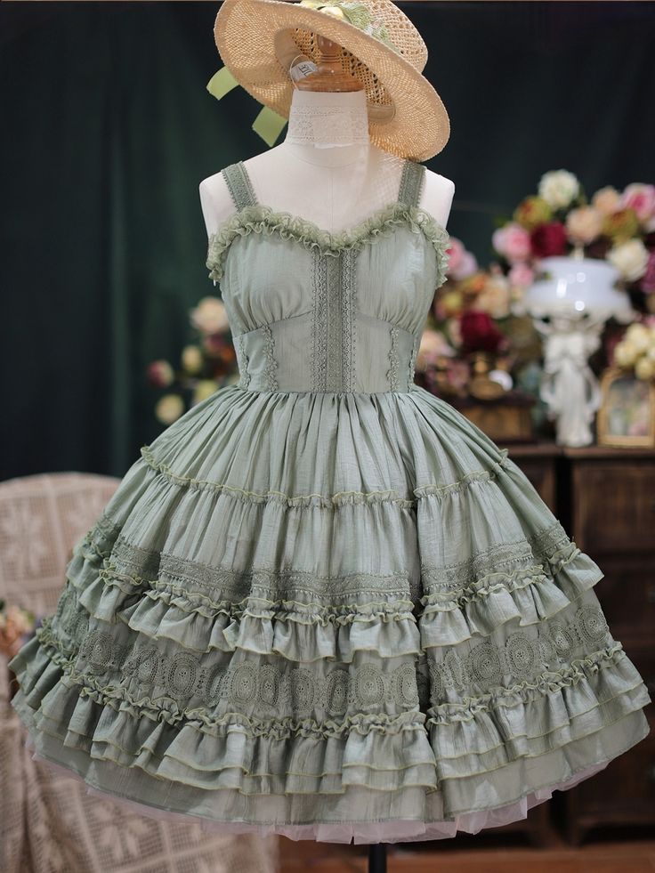 a sage green lolita fashion dress is displayed on a mannequin. the dress features a sweetheart neckline, delicate lace detailing, and multiple layers of ruffles on the skirt. the mannequin is wearing a straw hat, and there are flowers and a vintagestyle lamp in the background.