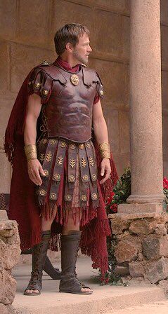 a man dressed as a roman centurion stands in front of a stone wall. he is wearing a leather breastplate, a segmented leather skirt, a red cloak, and leather sandals. his armor has gold accents. he has short brown hair and a serious expression.  the setting suggests an ancient roman environment.