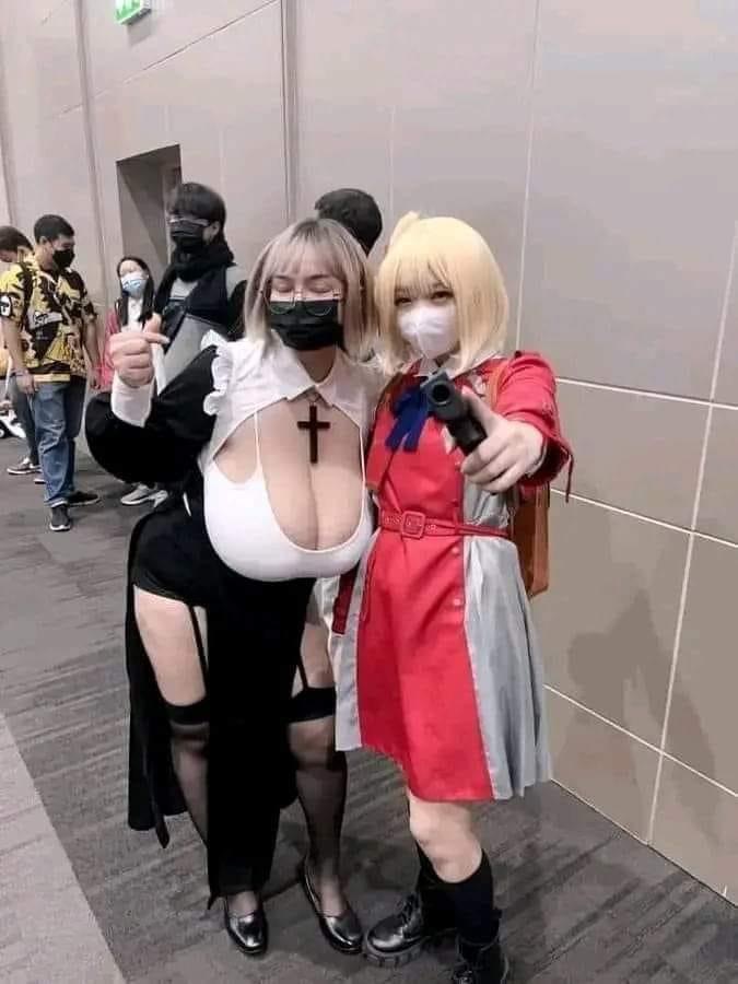 two women cosplaying as anime characters. the woman on the left is dressed as a nun with a black dress and a cross on her chest. the woman on the right is dressed in a red and grey dress and is pointing a gun at the camera. they are standing in front of a wall and a crowd of people. both are wearing face masks.