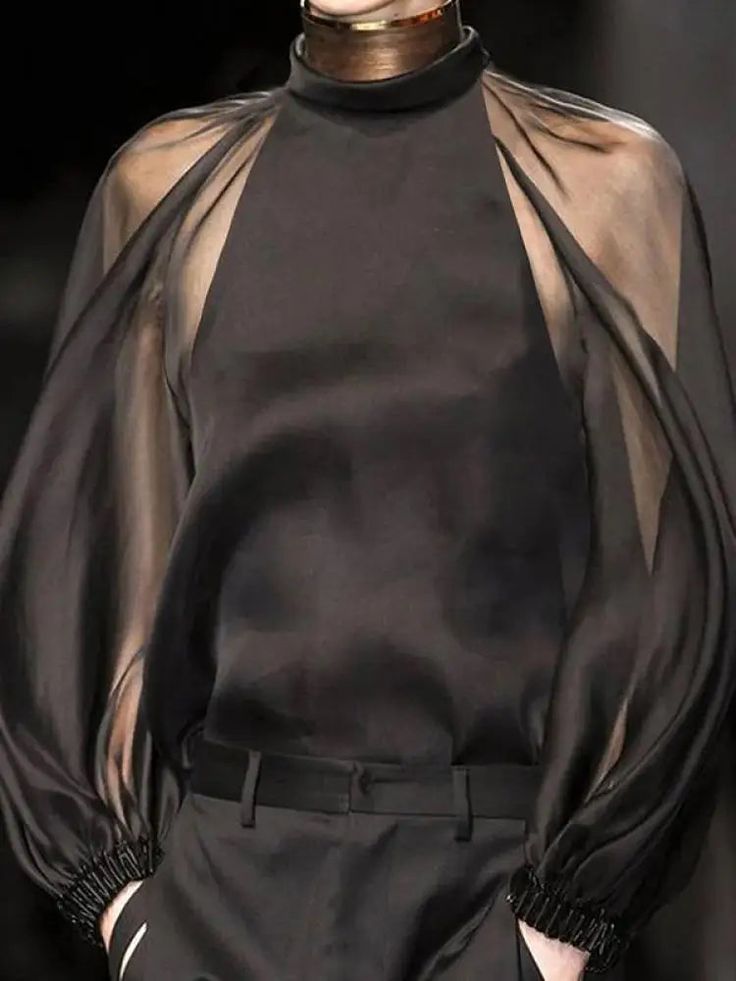 a woman in a black sheer blouse with a turtleneck, and black pants. she is wearing a gold collar. the woman is posing for a fashion shoot. the outfit is stylish and elegant. the picture is taken in a highend environment, and the lighting is soft and flattering.