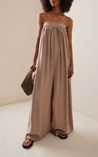a woman with dark brunette hair is standing in a studio with a beige background. she is wearing a widelegged, light beige jumpsuit. the jumpsuit has a square neckline and thin straps. she is holding a brown bag in her left hand and she is wearing black sandals with an embellishment on the straps. she is also wearing a necklace with a small heart pendant. her hair is long and styled straight with bangs. she is looking to the right. the photo is a medium shot, and she is posed in a relaxed, casual manner.