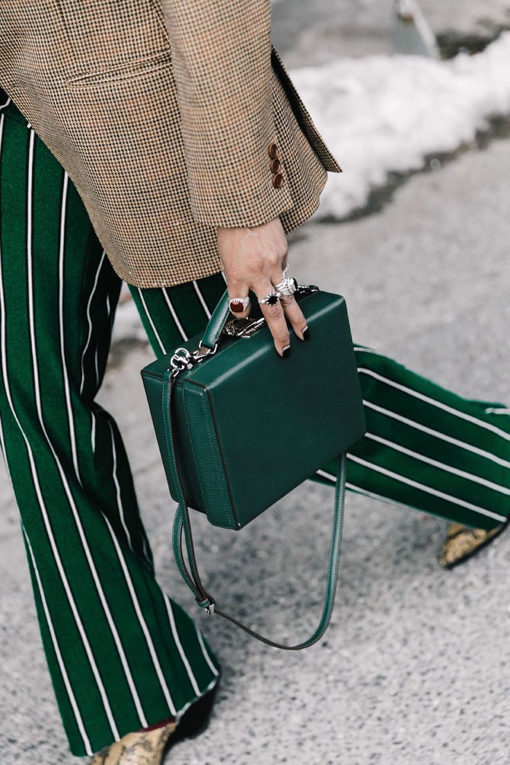 a woman is walking down a street, her outfit is a combination of a brown and beige checked blazer, green and white pinstripe pants, and a dark green box handbag. she's wearing several rings. the photo is taken from a medium shot, with a focus on the woman's attire and accessories. the background is blurry but shows a snowy or icy pavement. the overall style is chic and elegant.
