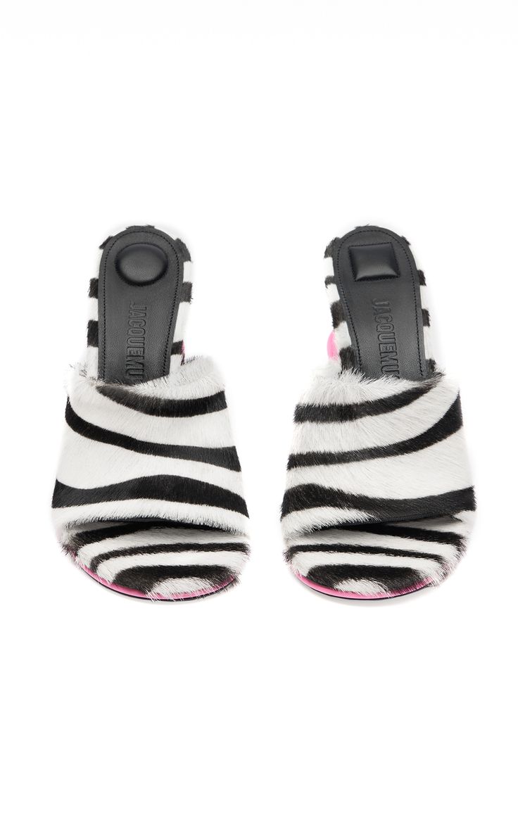 a pair of jacquemus women's slides in a zebra haircalf print. the sandals have a black and white zebra pattern upper with a pink sole.  they are styled as mules (slippers) with open toes and a slightly elevated heel. the jacquemus branding is visible on the insole. the image shows the shoes from above, against a white background.