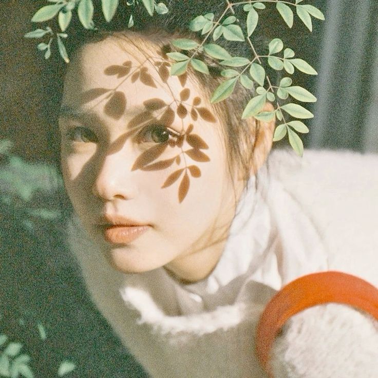 a closeup portrait of a young woman, likely asian, with her face partially obscured by green leaves. the leaves create a shadow pattern across her face, adding an ethereal and romantic touch to the image. the woman's eyes are looking directly at the camera, and her expression is soft and gentle. the background is blurred, highlighting the subject and creating a sense of intimacy. the image conveys a sense of peace, beauty, and natural elegance. the lighting is soft and warm, adding to the dreamlike atmosphere.