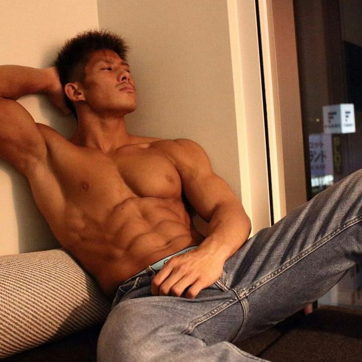 a muscular man with a six pack is sitting on a couch, shirtless, wearing jeans. he has one arm resting behind him and is looking up. the photo is taken from a low angle.  there is a window behind him and we can see city lights through the window.