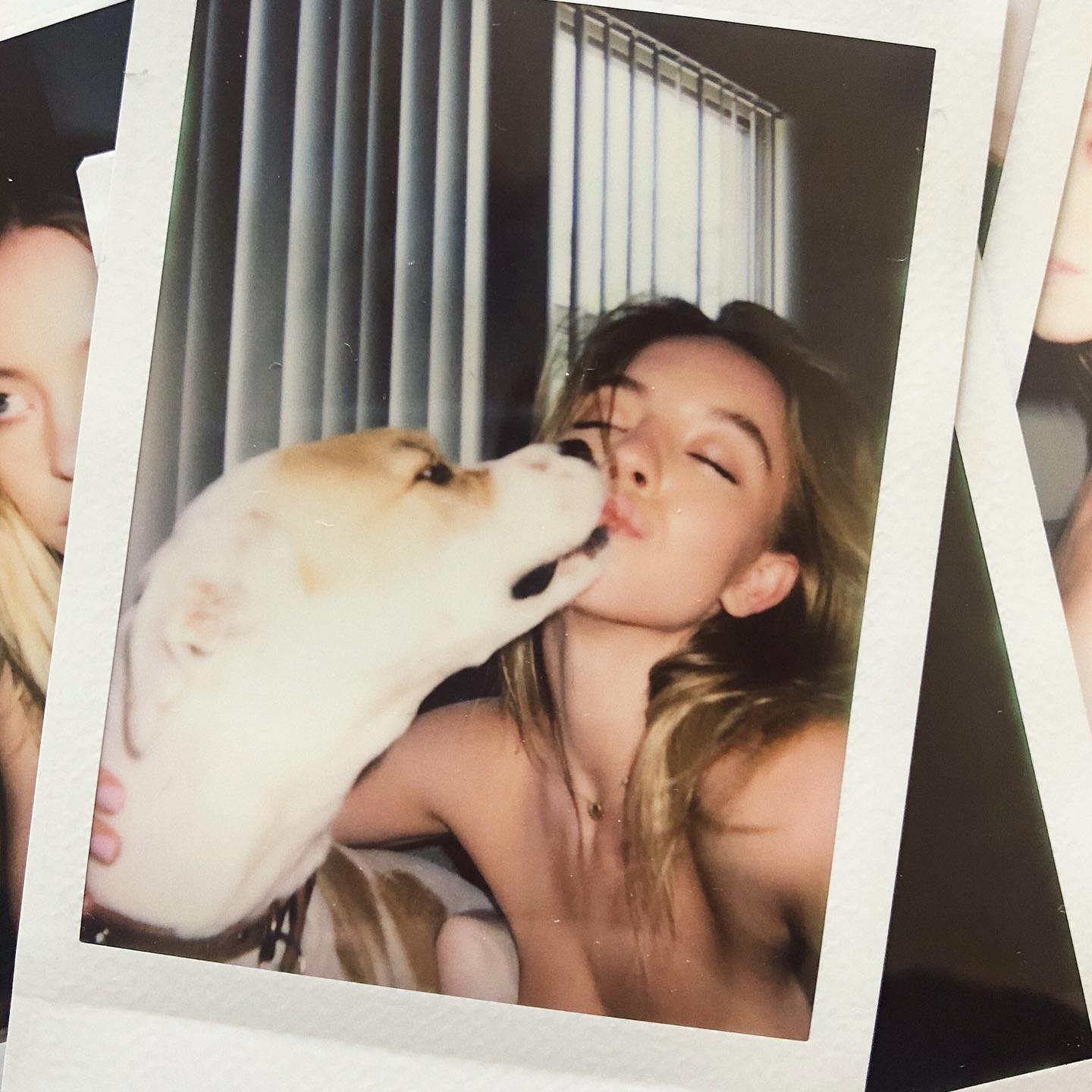 the image is a polaroidstyle photograph showing a young blonde woman with bare shoulders kissing a lightcolored dog. the woman's eyes are closed, and she appears to be enjoying the moment.  the background is slightly out of focus but shows a window with curtains. the overall feel is intimate, casual and slightly nostalgic due to the polaroid format and the soft focus.  other polaroid photos are partially visible in the corners.