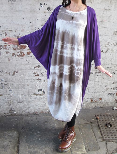 a woman stands in front of a brick wall and sidewalk, wearing a casual outfit. she is wearing a tiedye dress under a long purple cardigan. she also wears black leggings and brown leather boots. a silver necklace with a pendant is visible around her neck. her arms are outstretched.