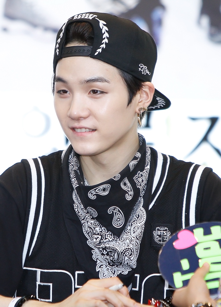 a headandshoulders shot of a man wearing a black baseball cap with white leaflike decorations, along with earrings and a black bandana with white paisley patterns around his neck. he's also wearing a black shirt or jacket with white stripes.