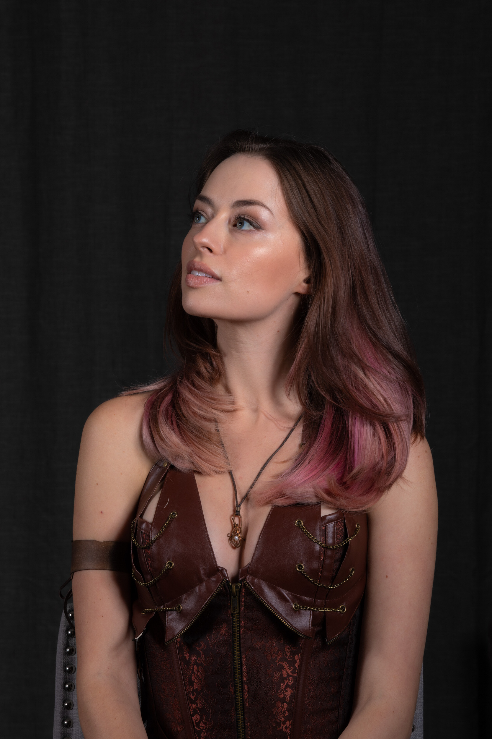a woman with pink and brown hair and wearing a brown leather corset  is photographed against a black backdrop. she gazes offcamera to the left, with her head turned slightly upward. the image is a studio portrait, lit with soft lighting.