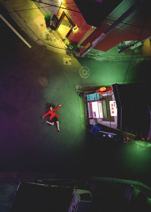 an aerial view of a woman lying on a dark street at night in a japanese city. she is wearing a red jacket and lying on her back with her arms outstretched. the street is empty and deserted, and the buildings around her are lit up. the scene has a dark and mysterious feel to it, and it could be interpreted as a crime scene.