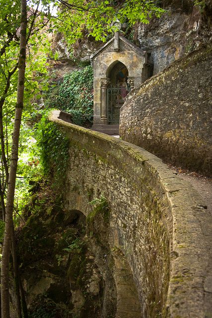 a stone wall and path lead to a small stone shrine nestled into a hillside. lush green vegetation surrounds the scene, and a bridge with an archway supports the pathway. trees and nature are prominent features in the landscape.