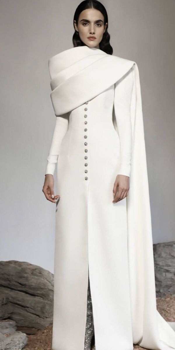 a full shot of a model posing in a long, white dress with a dramatic capelike overlay. the dress features a high neck, long sleeves, and a buttondown front.  the model is posed against a backdrop of rocks and a lightcolored background. the overall style is minimalist and elegant. the dress is a modern, sophisticated design.