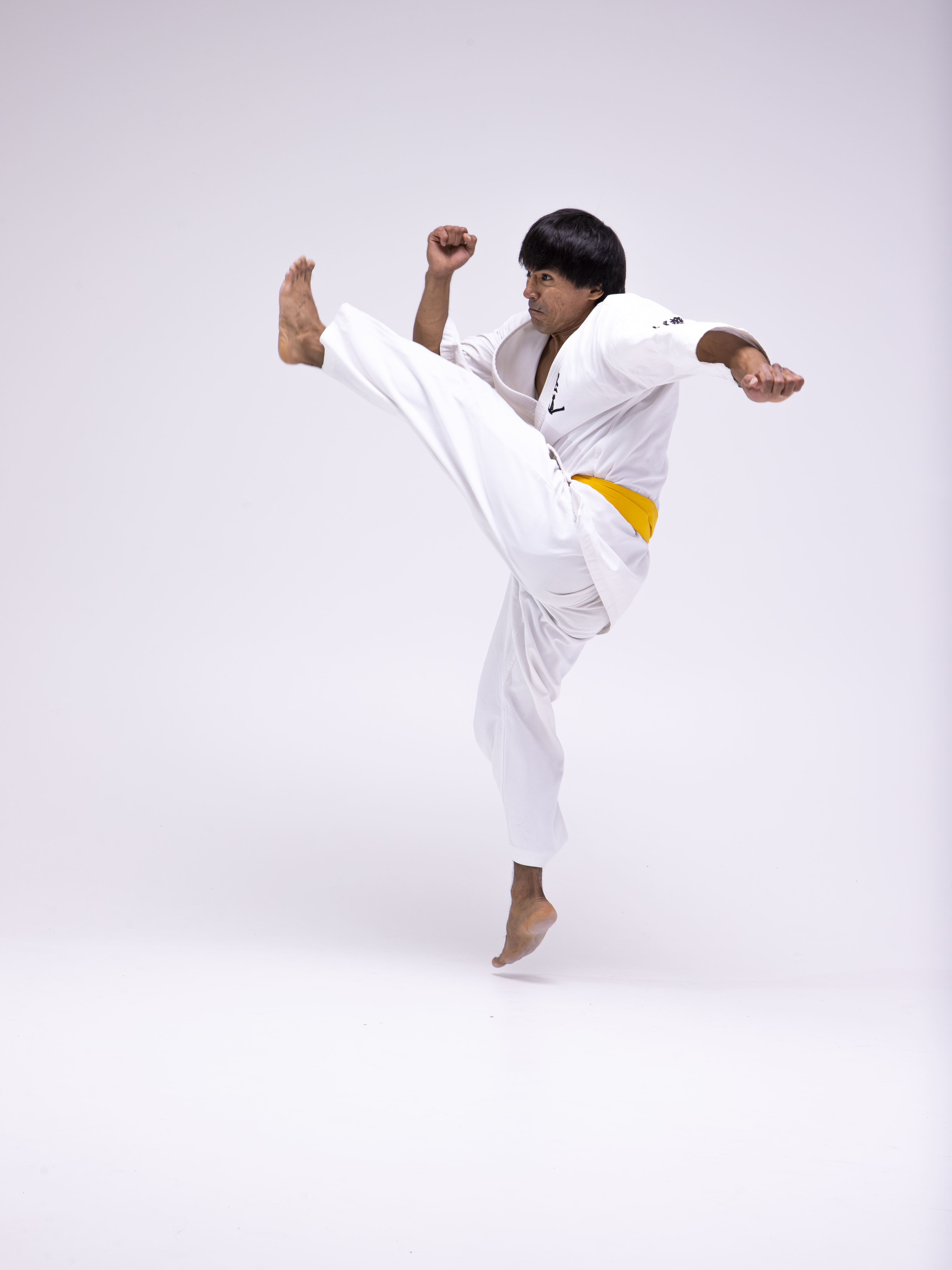 a man in a white karate uniform with a yellow belt is performing a high kick in midair against a white background. he is barefoot, and his arms are positioned for balance or defense.