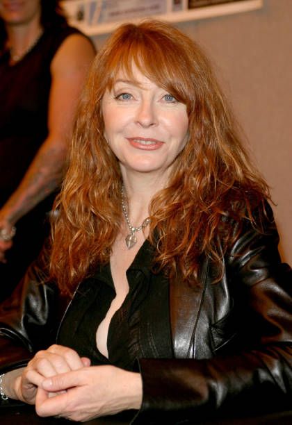 a young woman with long red hair and a leather jacket is smiling and looking at the camera. she is wearing a black top with a silver chain necklace and is posing for a portrait.